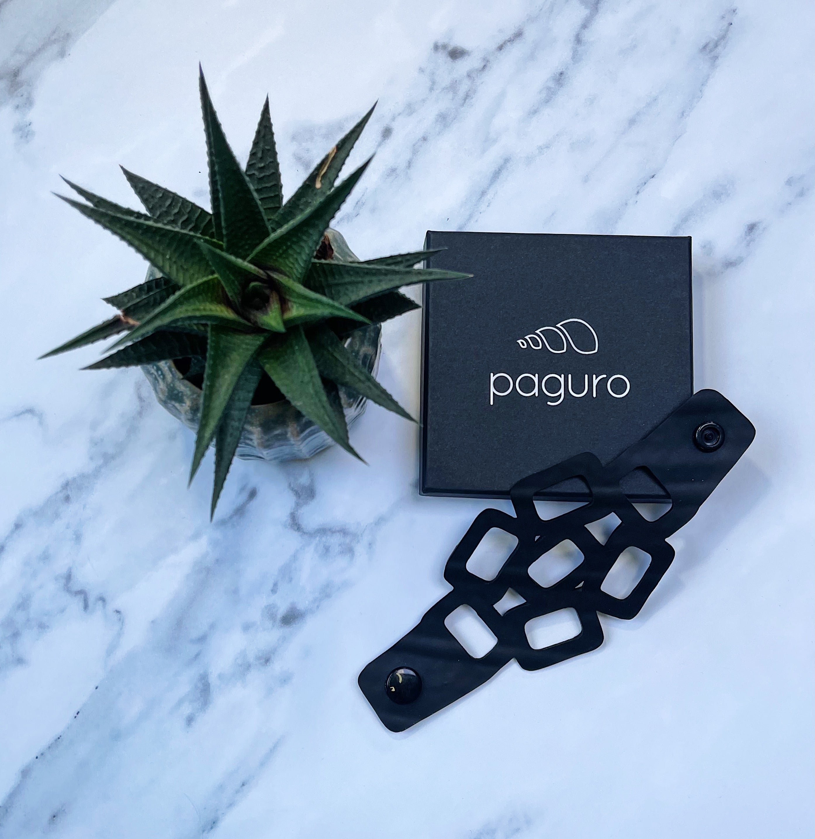 Paguro Upcycle eco friendly chain bracelet in black displayed next to plant and branded box on marble surface