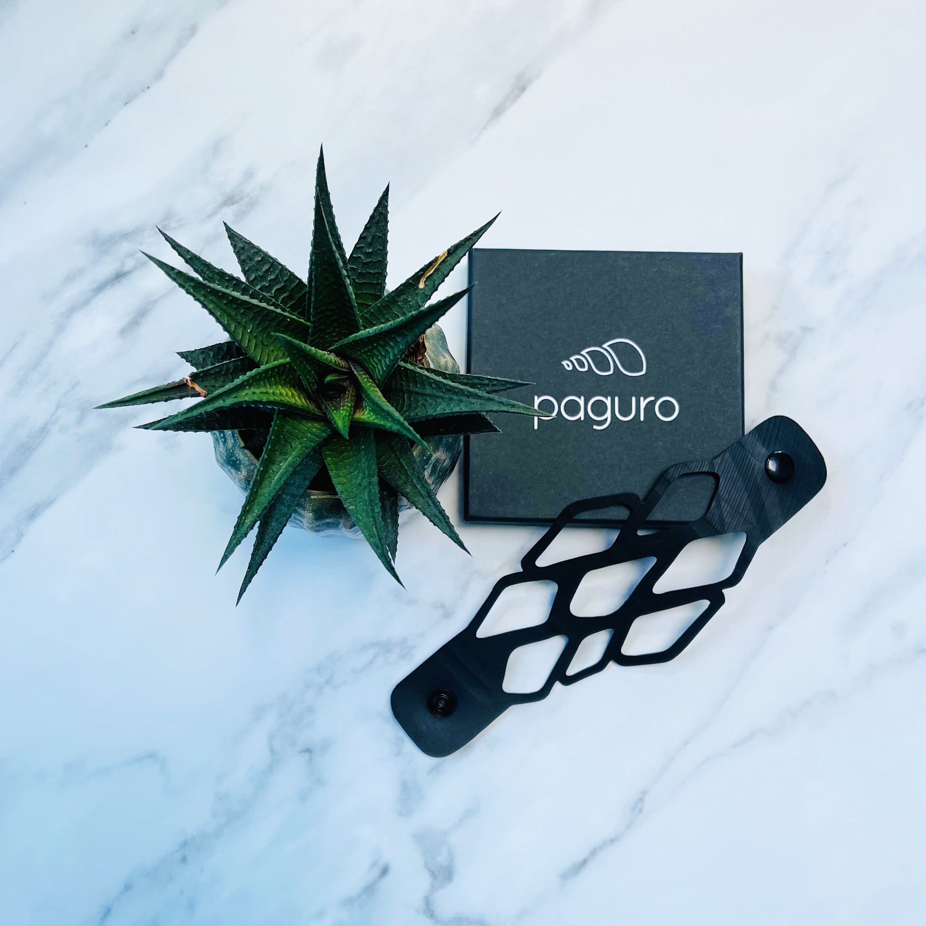 Paguro Upcycle black recycled rubber bracelet with geometric cutouts displayed next to green succulent plant and black Paguro box