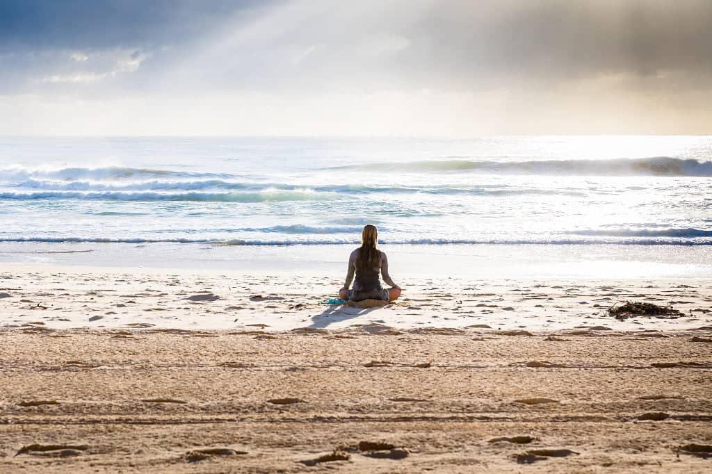 Woman practising meditation on a beach, embracing holistic living benefits in a serene environment