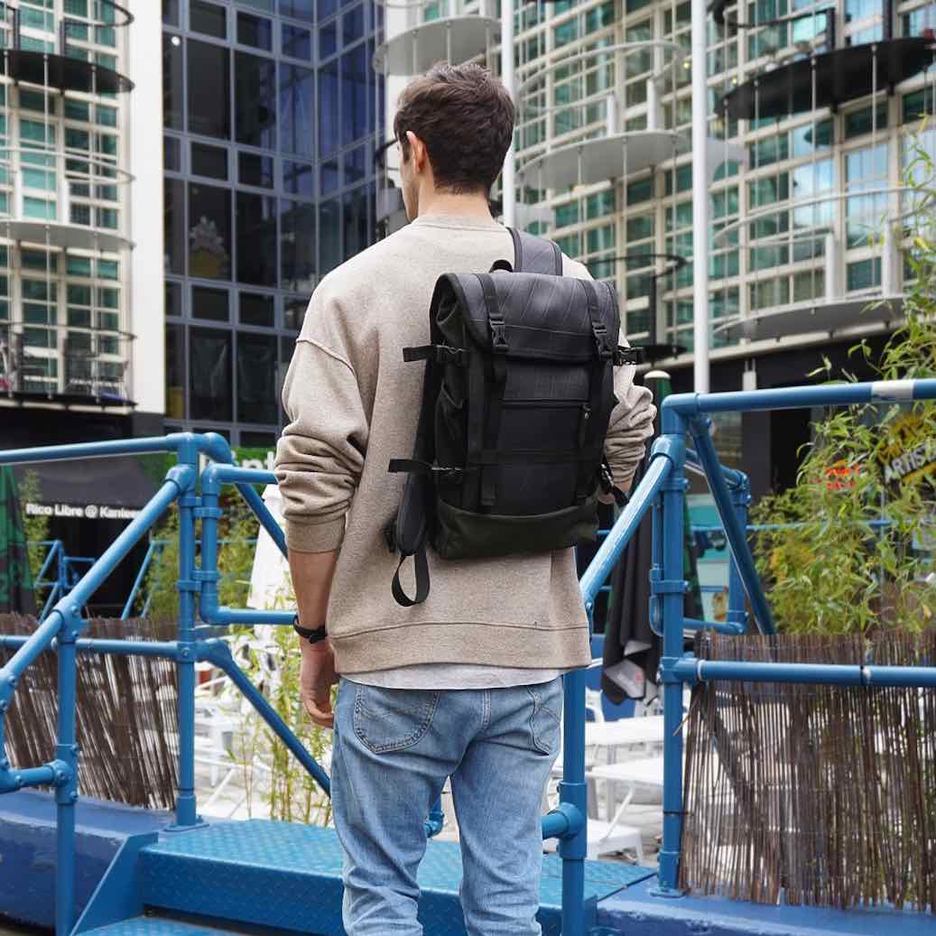 Person with black backpack walking on blue bridge near modern buildings.