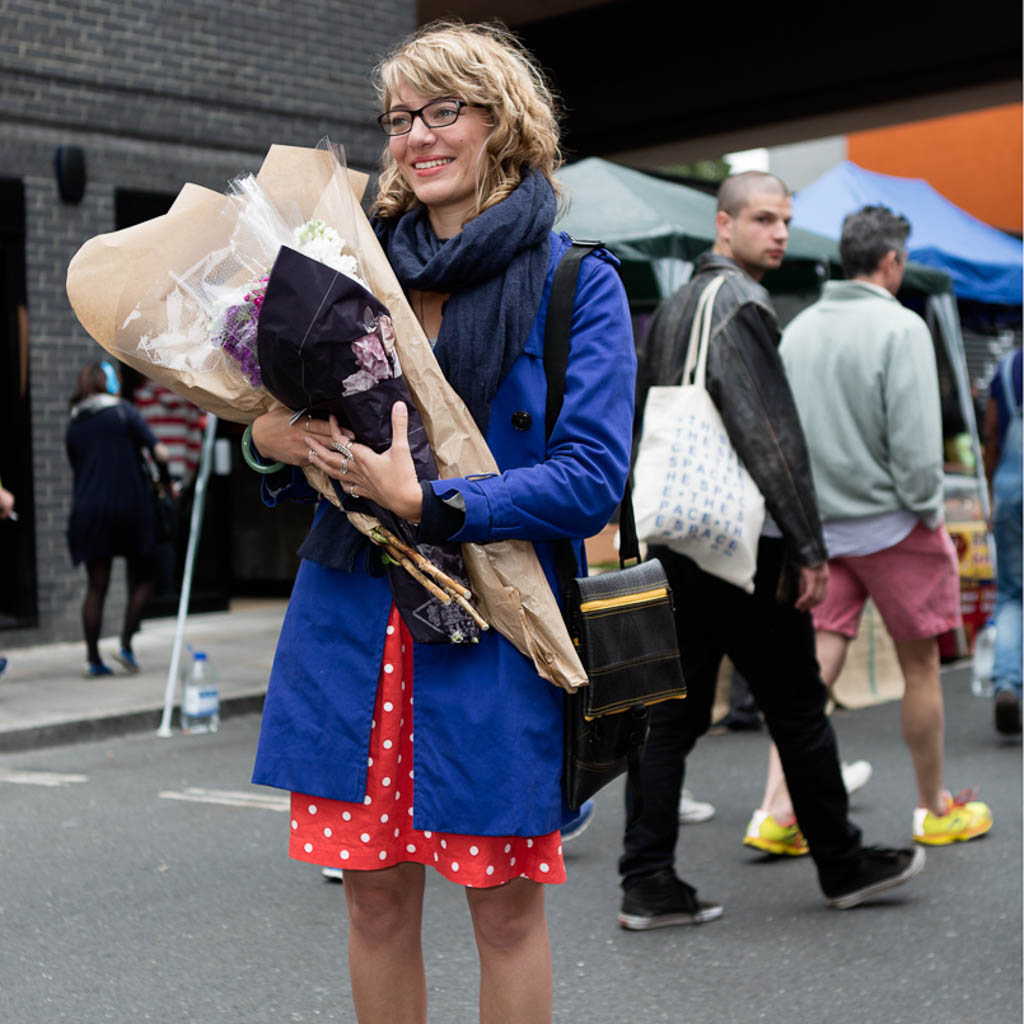 Woman wearing blue coat carrying recycled rubber crossbody bag holding a bouquet of flowers at outdoor market